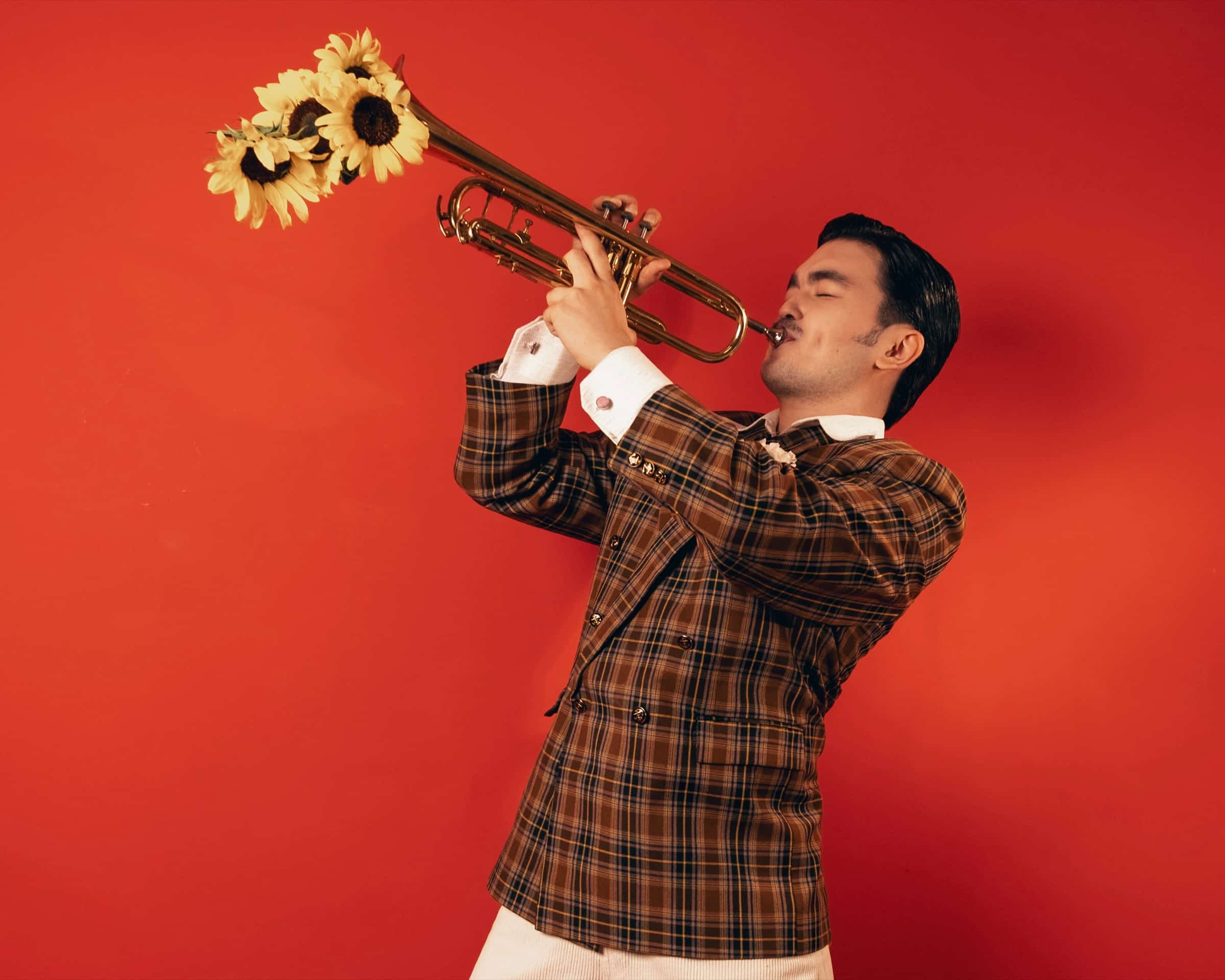 Almás playing a trumpet with sunflowers in the bell, against a red wall